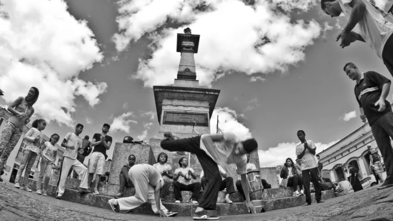 Jogo de capoeira em roda de rua, com capoeirista em movimento e público ao redor