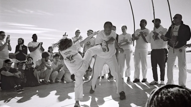 Dois capoeiristas jogam no centro de uma roda ao ar livre, cercados por músicos com berimbaus e público
