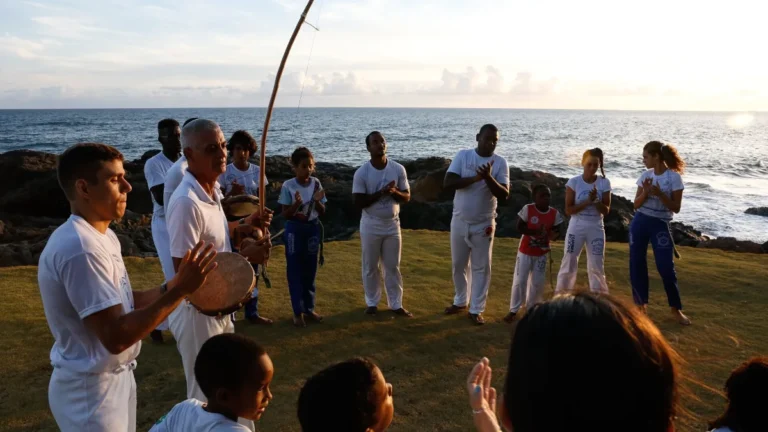 Roda de capoeira à beira-mar ao entardecer, com mestre tocando berimbau e alunos batendo palmas